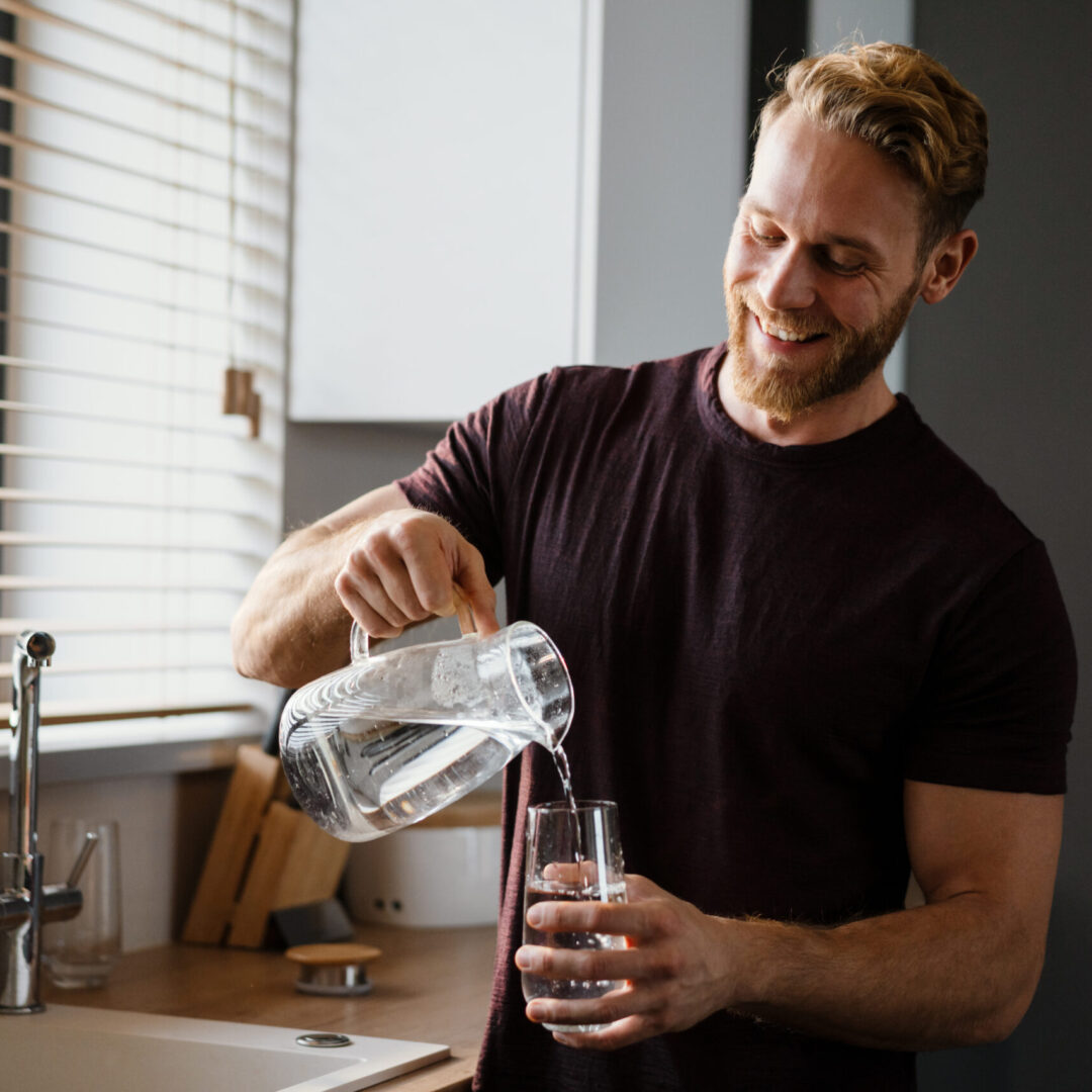 Attractive young man pouring water from a juf in a glass while standing at the kitchen sink